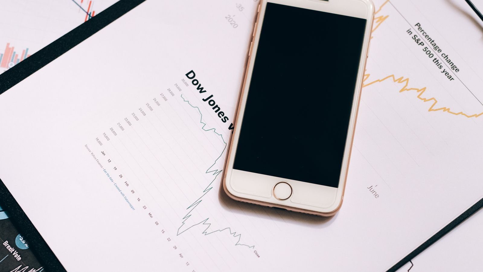 A smartphone and printed market charts illustrating a comparison of investment holdings and price information