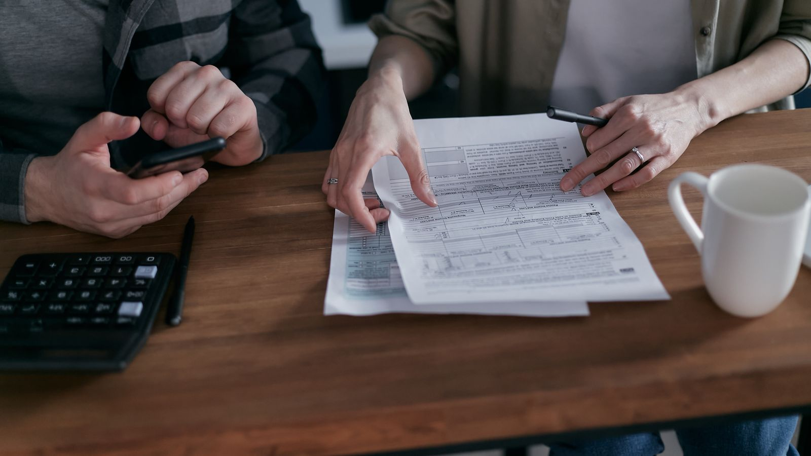 Two people checking payment details using a phone, documents, and a calculator